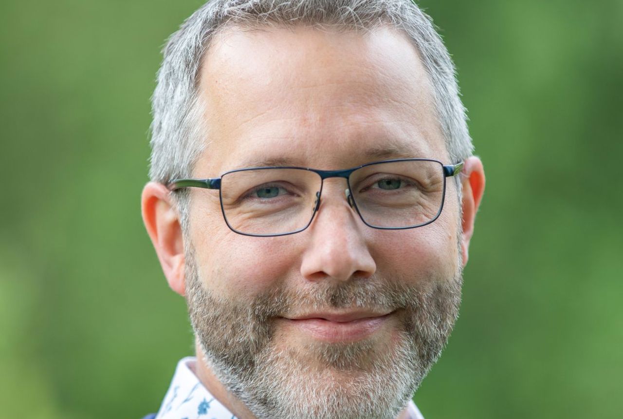 A man in glasses and a blue jumper smiles at the camera