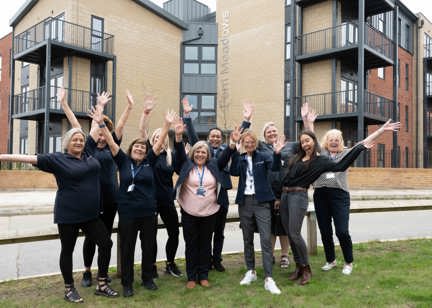  A group of Housing 21 colleagues standing on grass positioned at the front entrance outside Fern Meadows with their arms up cheering