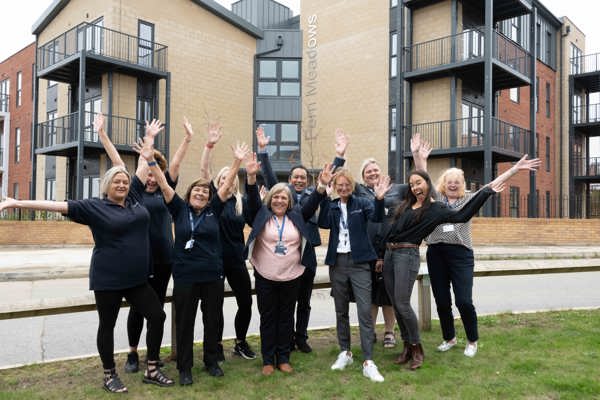 A group of Housing 21 colleagues standing on grass positioned at the front entrance outside Fern Meadows with their arms up cheering