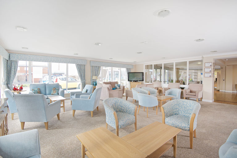 Residents' communal lounge with soft seating and tables in light blue colour scheme