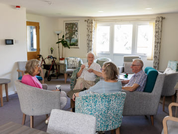 Residents sitting drinking tea and socialising in a communal lounge with purple carpet, teal and grey arm chairs, with coffee tables.