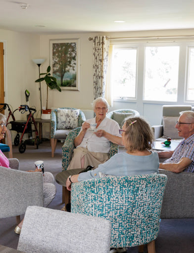 Residents sitting drinking tea and socialising in a communal lounge with purple carpet, teal and grey arm chairs, with coffee tables. 