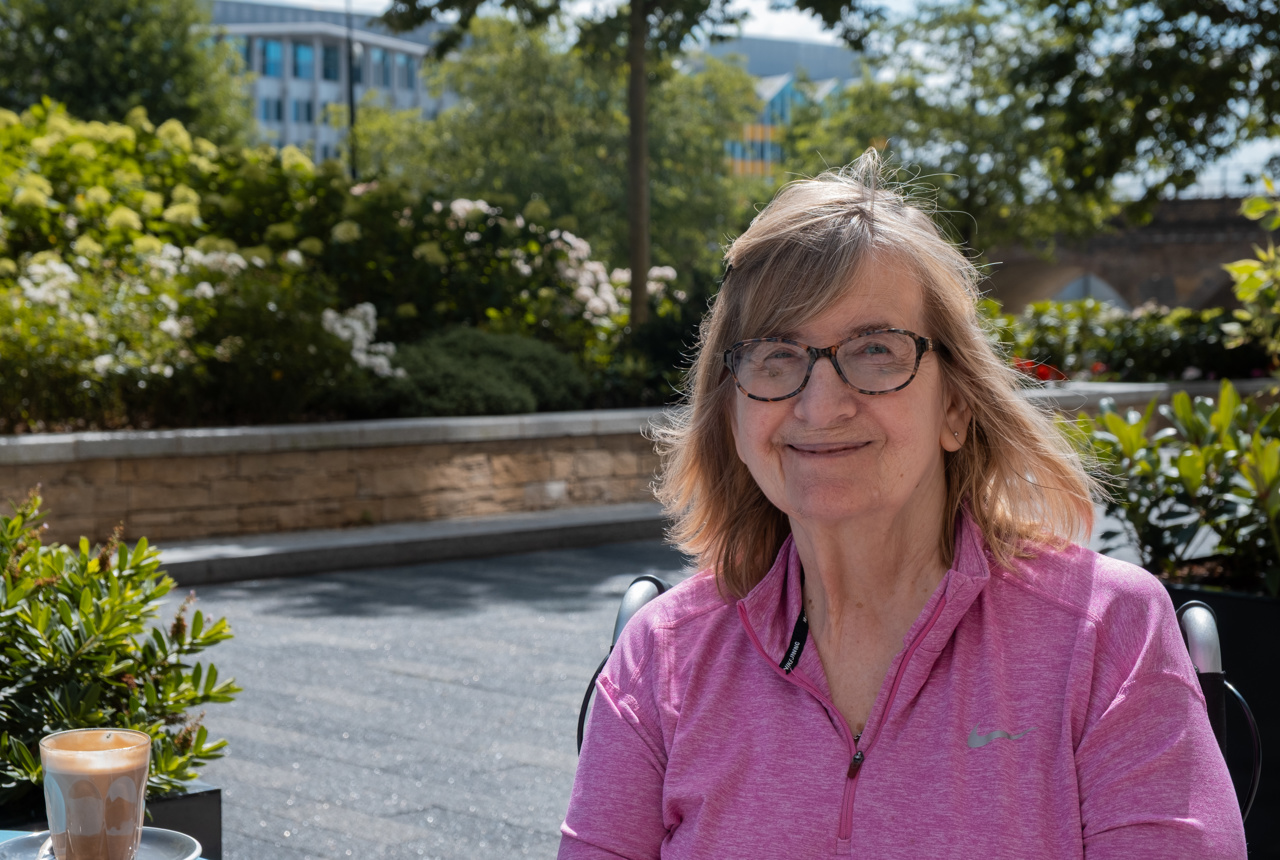 Resident Janet smiling sitting in the garden in a wheelchair wearing a pink sports jacket