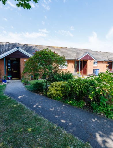 A row of bungalows set behind hedging with a footpath to the front 