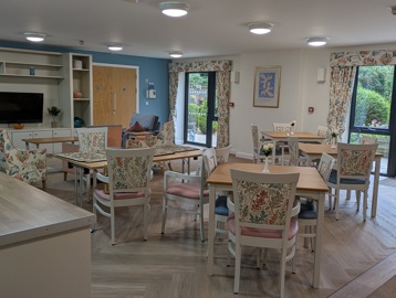 A communal dining area, dining chairs with floral designs pulled up at wooden square tables. There's a television set on the wall.