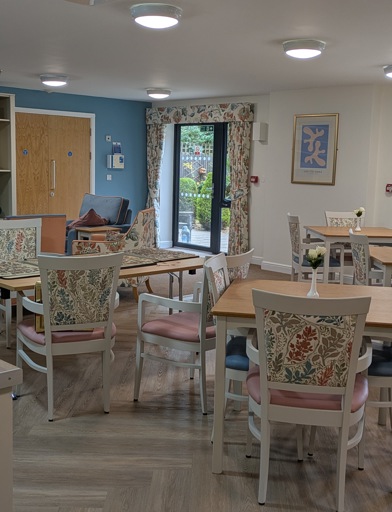 A communal dining area, dining chairs with floral designs pulled up at wooden square tables. There's a television set on the wall. 