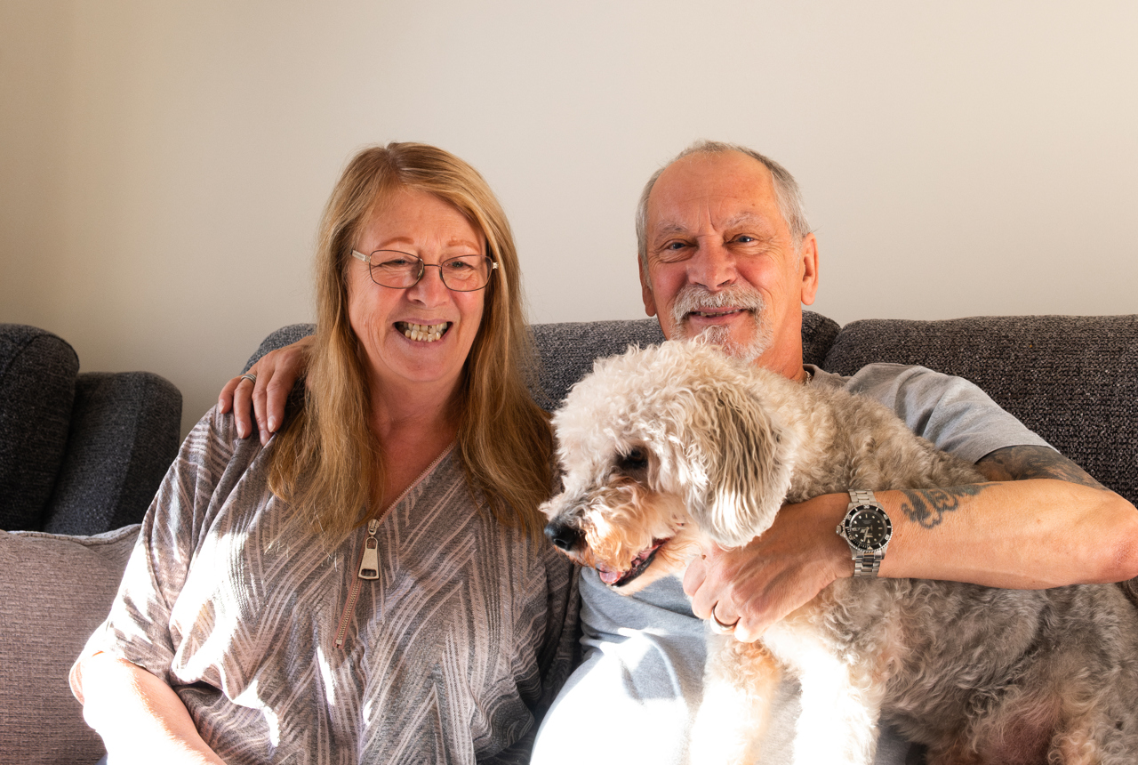 A smiling couple in their apartment with their pet dog sat beside them on the sofa