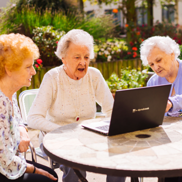 Three ladies gather around a laptop whilst sitting in a garden 