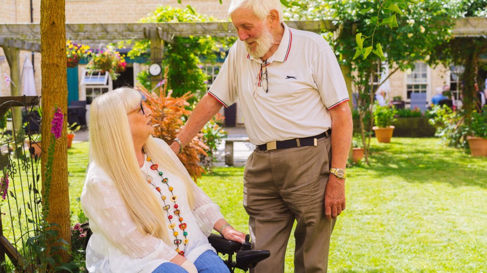 Resident in a wheelchair in a communal garden on a sunny day looking up to and smiling at a friend
