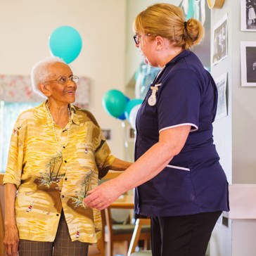 An Extra Care resident and care worker talking in a communal lounge with chairs and balloons around the room