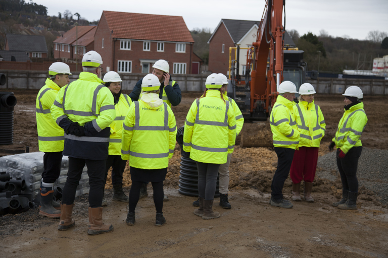 Housing 21 employees along with key representatives stood outside on muddy ground, wearing high vis jackets and hard hats with construction equipment. 