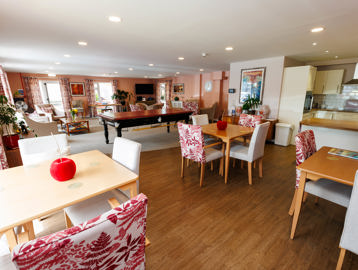 Communal lounge with a beige and red colour theme with dining chairs and tables and kitchen area