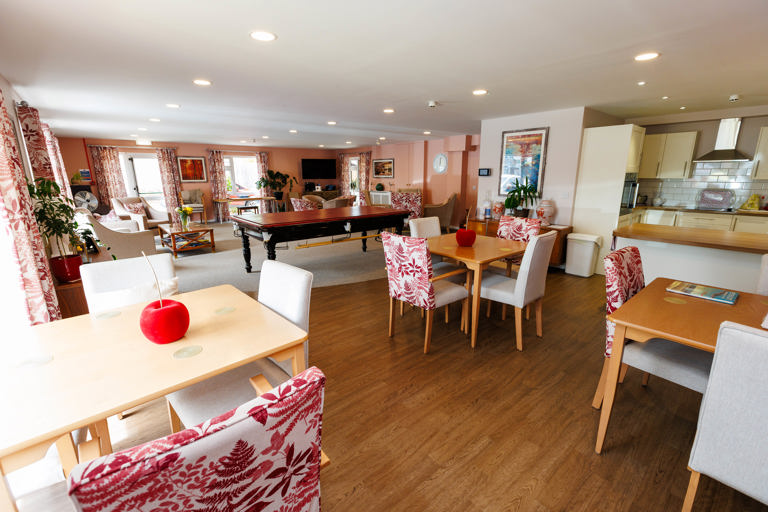 Communal lounge with a beige and red colour theme with dining chairs and tables and kitchen area