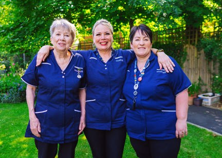 Three Care Workers wearing blue uniforms stood together in communal garden smiling into camera