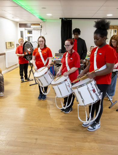 A children's marching band, three drummers lead, behind them a child plays keyboard, one on a clarinet and others using trumpets. 