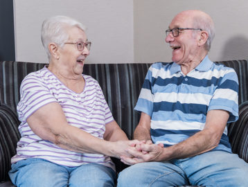 A couple facing each other and laughing on a sofa whilst holding hands 