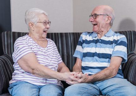 A couple facing each other and laughing on a sofa whilst holding hands 