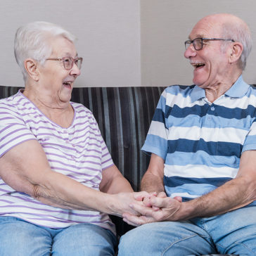 A couple facing each other and laughing on a sofa whilst holding hands 
