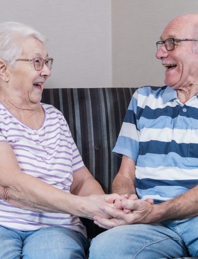A couple facing each other and laughing on a sofa whilst holding hands