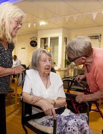 Lady in salmon pick top leans over to speak to a lady in a wheelchair 