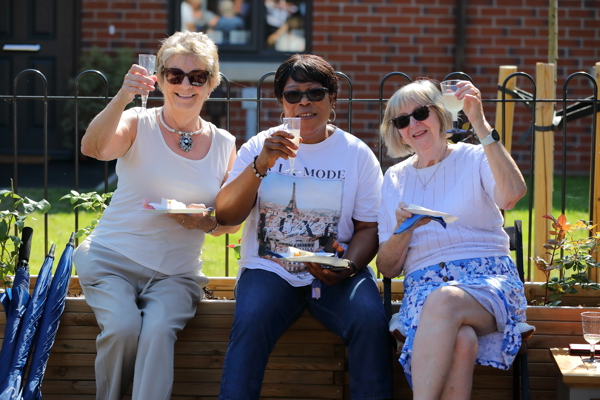 Three ladies smile whilst sitting on a flower boarder and raise a glass of prosecco to the camera