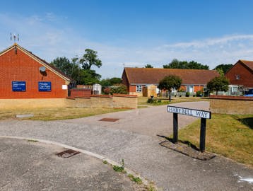 Entrance driveway to the scheme with bungalows, landscaped communal gardens and welcome signs.