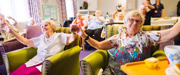 A group of residents sat in armchairs holding hands in the air and waving and smiling at the camera