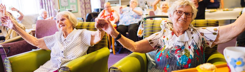 A group of residents sat in armchairs holding hands in the air and waving and smiling at the camera 