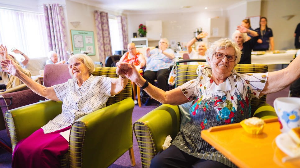 A group of residents sat in armchairs holding hands in the air and waving and smiling at the camera