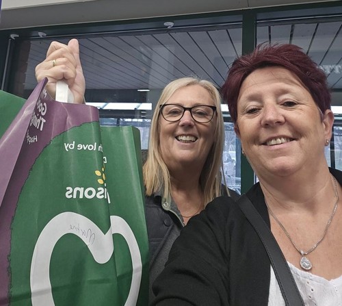 A Housing 21 employee and a Morrisons employee smiling and holding up a Morrisons bag full of shopping.