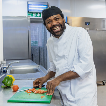 A chef wearing all whites smiles at the camera as he chops vegetables in the kitchen of an Extra Care scheme