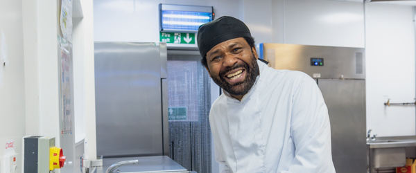 A chef wearing all whites smiles at the camera as he chops vegetables in the kitchen of an Extra Care scheme