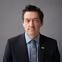 An image of a man wearing a dark suit with a light blue shirt and patterned tie 