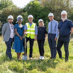 Housing 21 employees standing outside on grass wearing hard hats and holding a spade. 