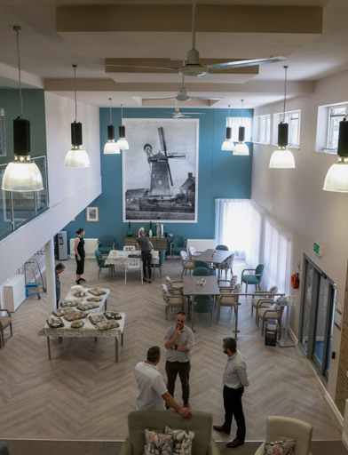 An ariel shot taken from a balcony showing a communal dining room and lounge, decorated with blue paint, a large photo of a windmill, parquet flooring and a living wall 