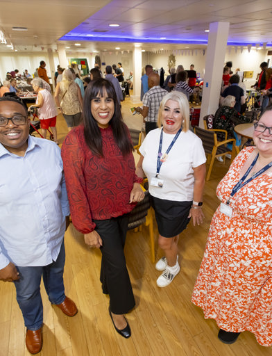 Three Housing 21 colleagues standing with MP Sureena Brackenridge with a room full of activity behind them including raffles and different stalls. 