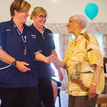 Two Care Workers in blue uniforms dancing in a communal lounge with a resident in bright yellow shirt