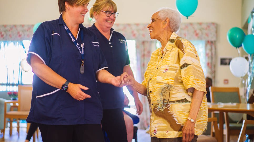 Two Care Workers in blue uniforms dancing in a communal lounge with a resident in bright yellow shirt