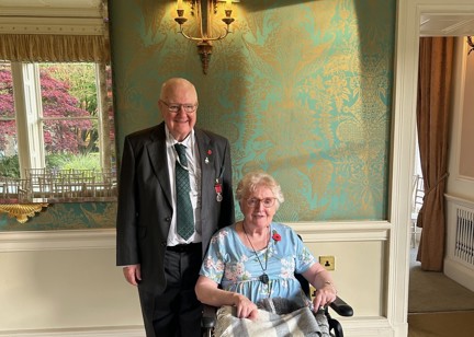 A couple smiling. A lady sits in a wheelchair wrapped in a blanket. A man stands behind, suited.