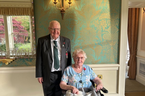 A couple smiling. A lady sits in a wheelchair wrapped in a blanket. A man stands behind, suited.