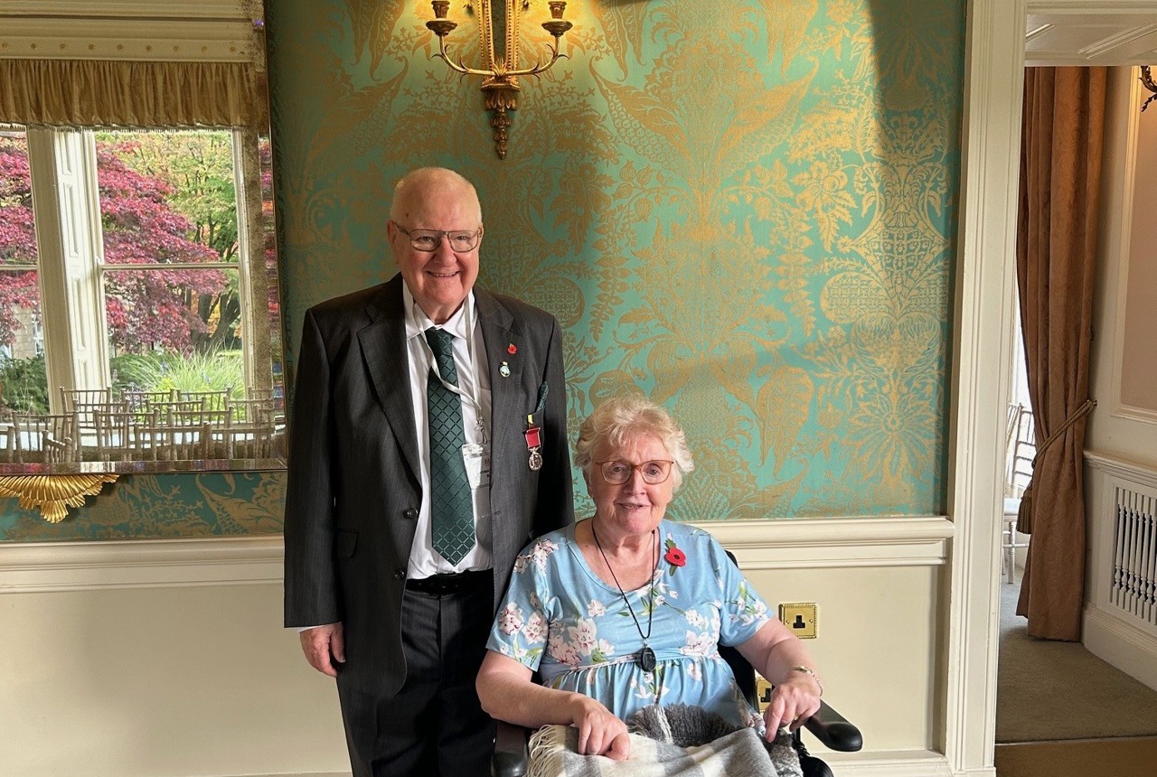 A couple smiling. A lady sits in a wheelchair wrapped in a blanket. A man stands behind, suited.