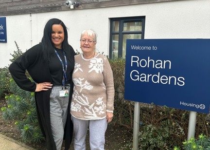 A Housing 21 employee and resident standing outside by the Rohan Gardens welcome sign. 