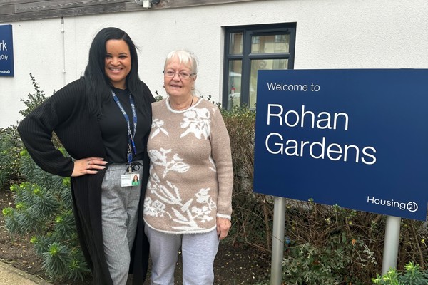 A Housing 21 employee and resident standing outside by the Rohan Gardens welcome sign. 