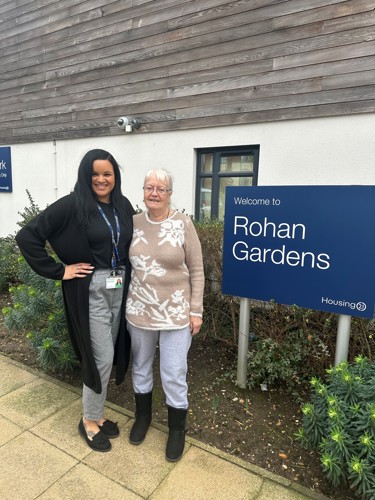 A Housing 21 employee and resident standing outside by the Rohan Gardens welcome sign.