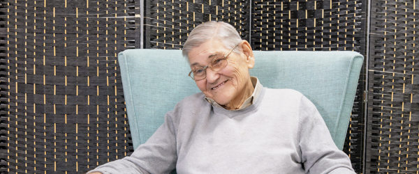 A resident sitting in an arm chair smiles at the camera