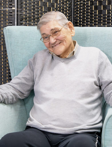 A resident sitting in an arm chair smiles at the camera 