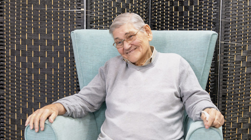 A resident sitting in an arm chair smiles at the camera