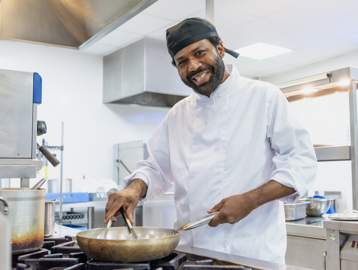 A chef wearing chef whites smiles whilst cooking at the hob with a frying pan