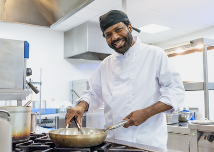 A chef wearing chef whites smiles whilst cooking at the hob with a frying pan