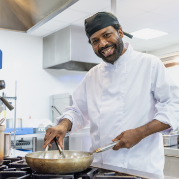 A chef wearing chef whites smiles whilst cooking at the hob with a frying pan